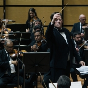 A imagem mostra uma apresenta&ccedil;&atilde;o de uma orquestra sinf&ocirc;nica em um palco de concertos. O fundo &eacute; formado por pain&eacute;is de madeira em tom claro, caracter&iacute;sticos de salas de espet&aacute;culo com tratamento ac&uacute;stico. No centro da cena est&aacute; o maestro, em p&eacute;, usando fraque preto, camisa branca e gravata-borboleta. Ele est&aacute; voltado para a orquestra, com o bra&ccedil;o direito elevado e a m&atilde;o aberta, conduzindo os m&uacute;sicos durante a execu&ccedil;&atilde;o da obra. &Agrave; sua frente h&aacute; um p&uacute;lpito com a partitura aberta. Ao redor do maestro est&atilde;o diversos integrantes da orquestra, vestidos com trajes formais pretos. Em primeiro plano e nas laterais aparecem violinistas e violistas sentados, segurando seus instrumentos e arcos em posi&ccedil;&atilde;o de execu&ccedil;&atilde;o, concentrados na leitura das partituras apoiadas em estantes pretas. Ao fundo, outros m&uacute;sicos tamb&eacute;m tocam instrumentos de cordas, compondo diferentes fileiras da orquestra.